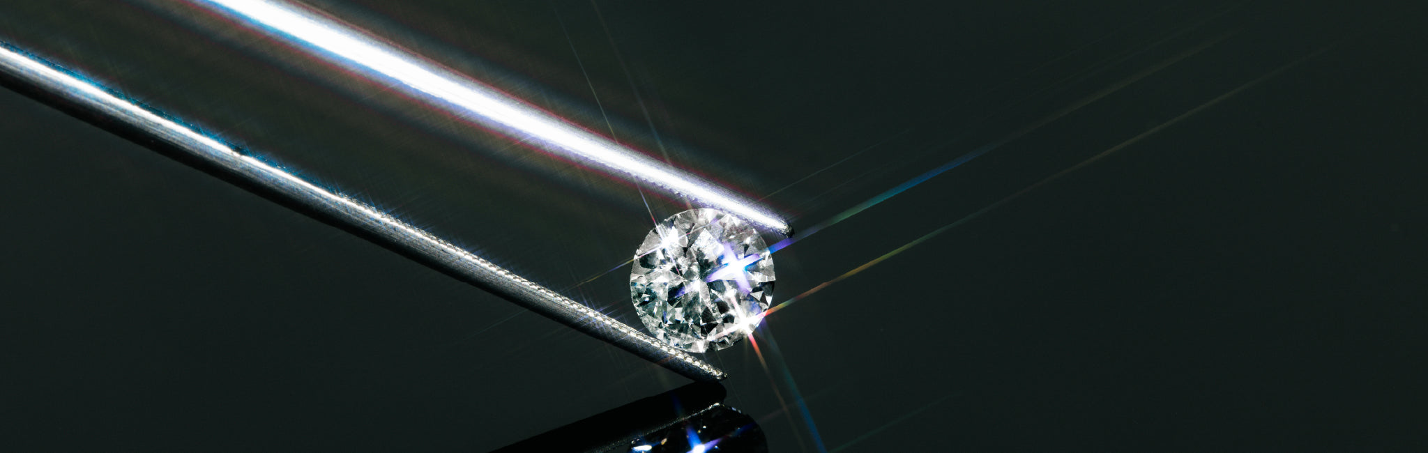 Brillantgeschliffener Diamant, gehalten von einer feinen Metallpinzette auf schwarzer, spiegelnder Fläche – Lichtreflexe erzeugen regenbogenartige Strahlen.