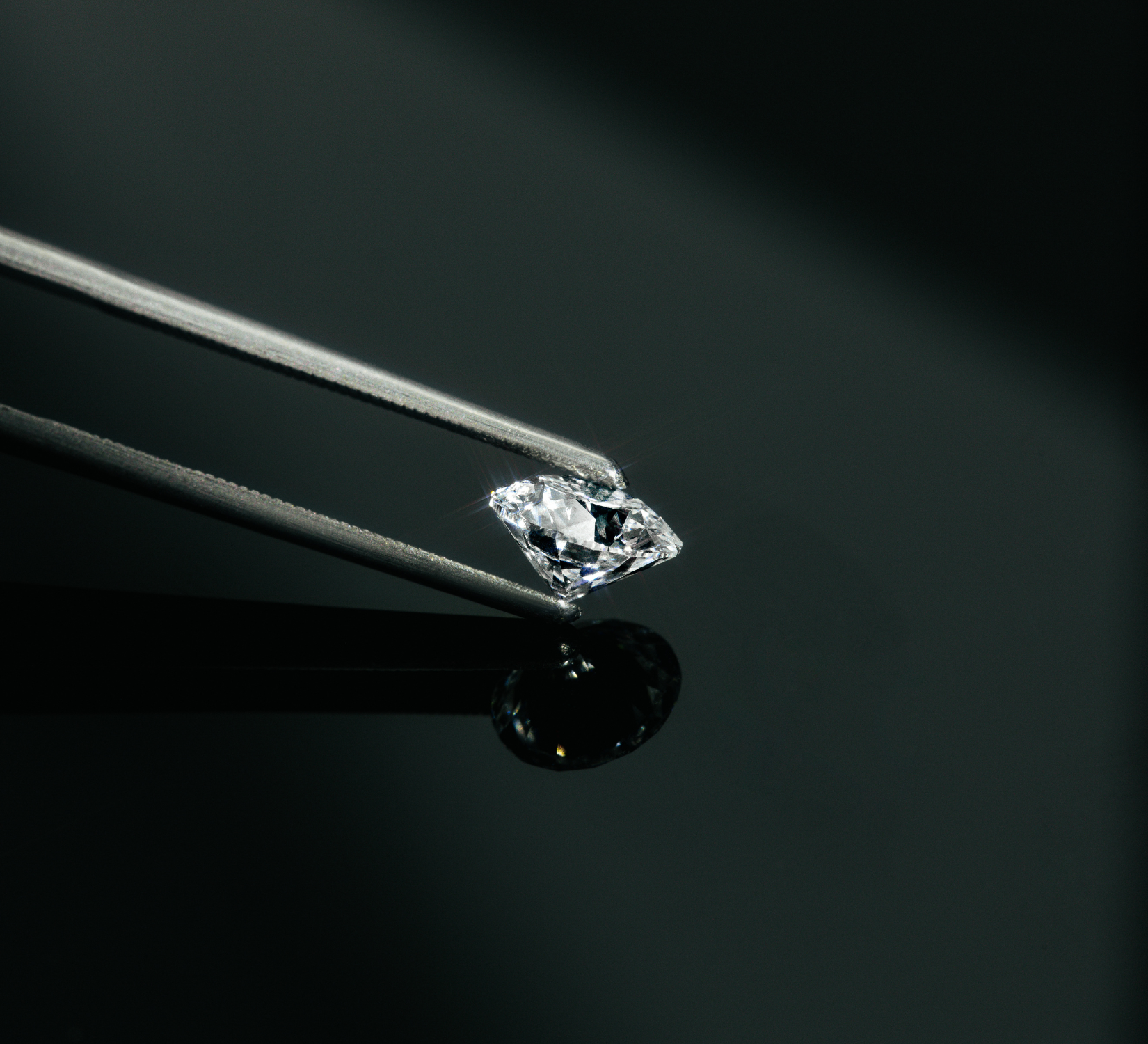 Brillantgeschliffener Diamant, gehalten von einer feinen Metallpinzette auf schwarzer, spiegelnder Fläche.