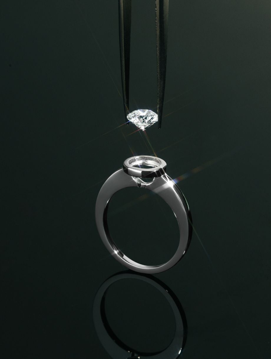 Nahaufnahme eines Solitärrings aus Weißgold mit offener Zargenfassung – ein Diamant wird mit einer Pinzette präzise über der Fassung gehalten, bereit zur Einfassung.