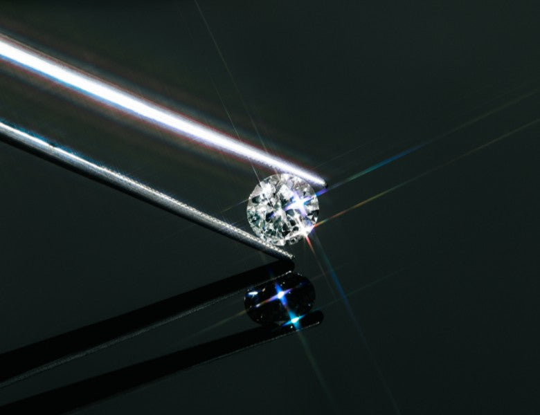 Brillantgeschliffener Diamant, gehalten von einer feinen Metallpinzette auf schwarzer, spiegelnder Fläche – Lichtreflexe erzeugen regenbogenartige Strahlen.