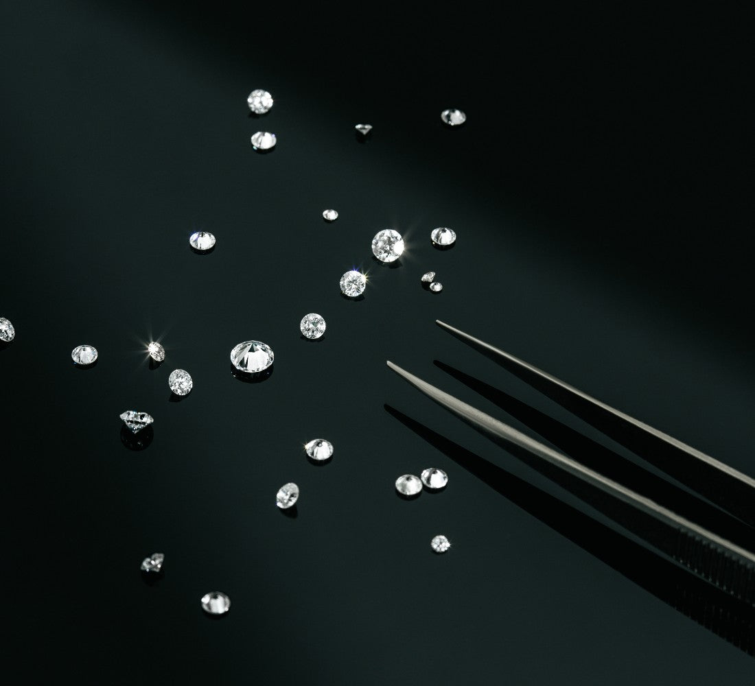 Verschieden große gezüchtete Diamanten liegen auf einem schwarzen Untergrund. Von der Seite kommt eine Zange ins Bild. 