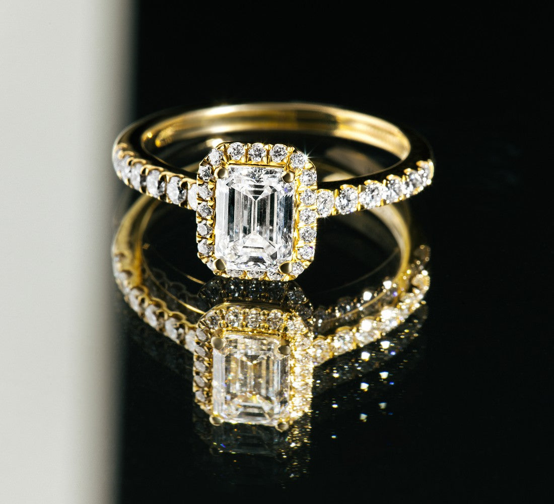 SIENNA Ring DS2067G aus Gelbgold mit rechteckigem Mittelstein im Emerald-Cut und umlaufenden Diamanten, reflektiert auf schwarzem Untergrund.